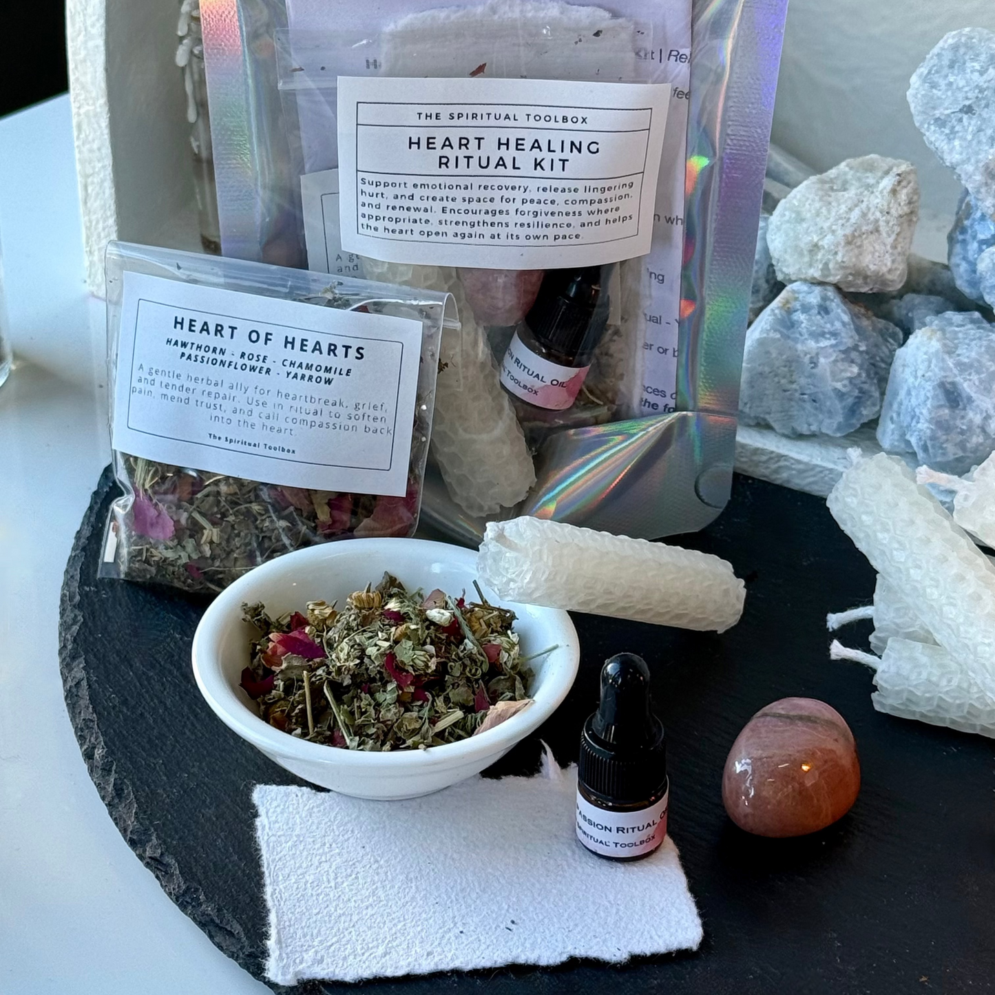 Heart Healing Ritual Kit by The Spiritual Toolbox, displayed on a black slate tray with crystals, white candles, a small bowl of dried herbs, and two small bottles.