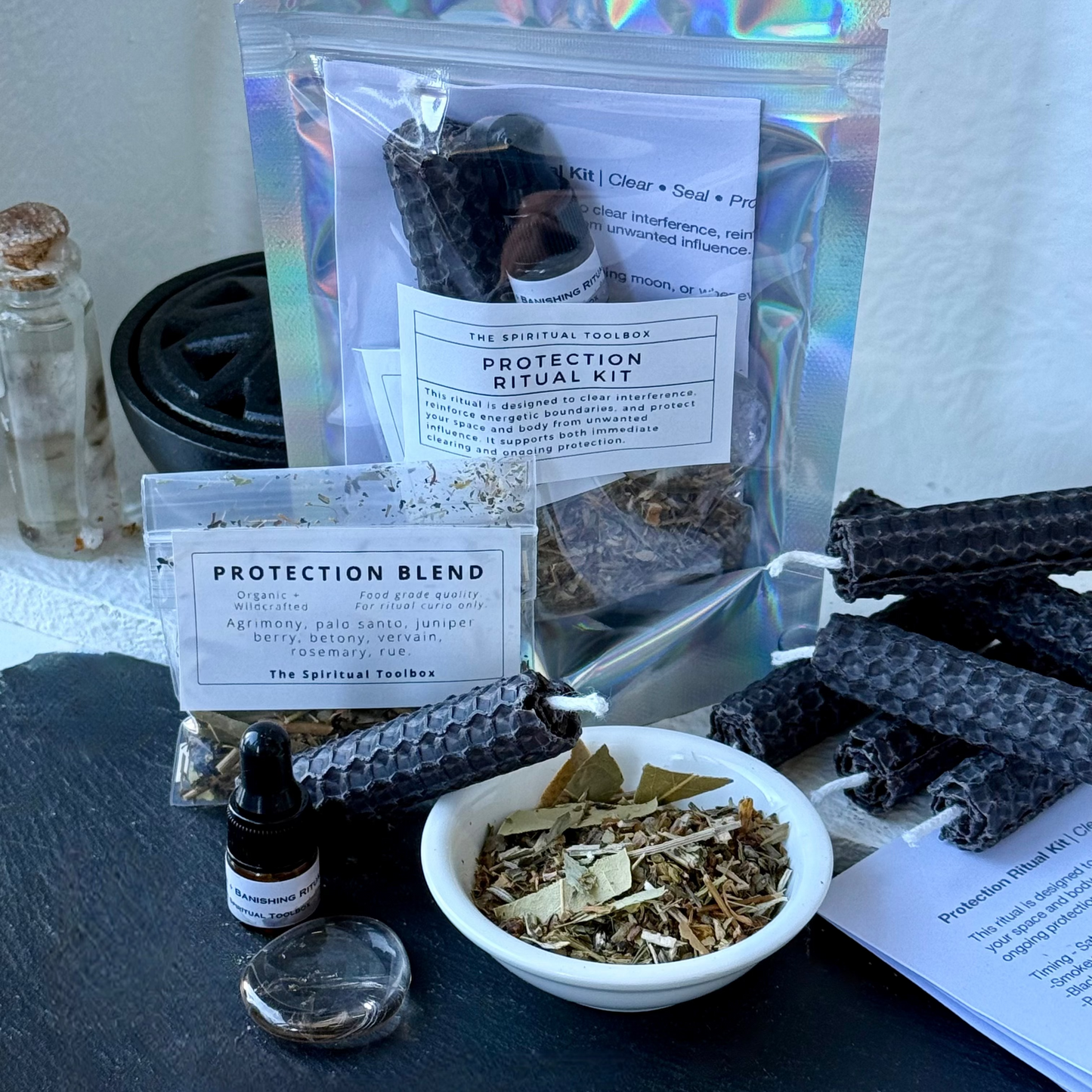 Protection Ritual Kit by The Spiritual Toolbox, featuring a holographic pouch of protection blend, a small dropper bottle, a white bowl of dried herbs, and several black beeswax candles on a dark slate surface.
