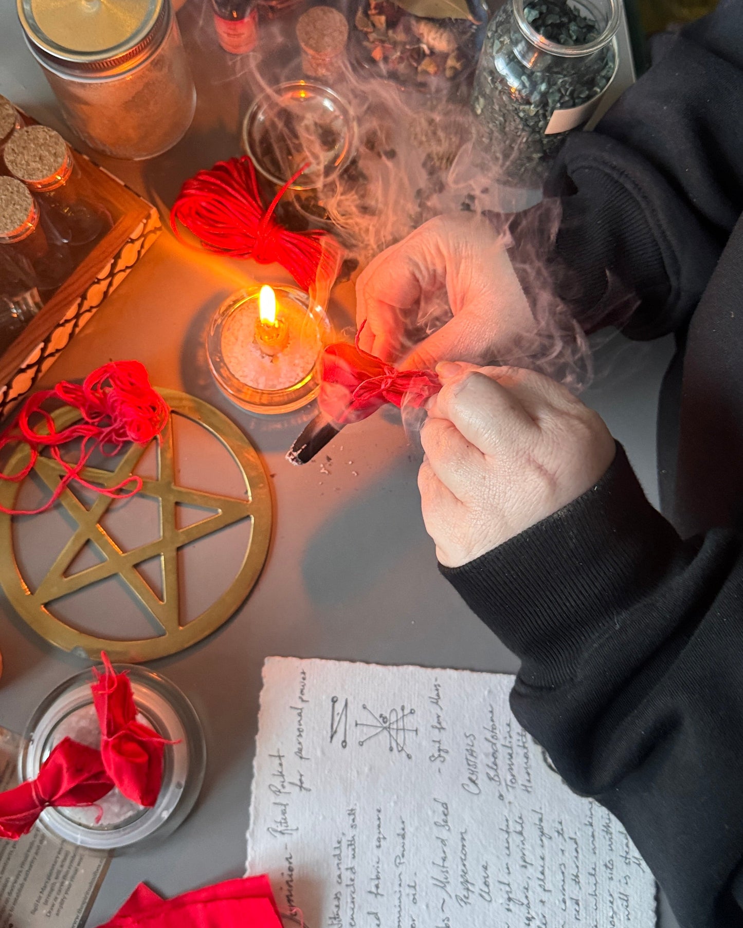 A person's hands are shown holding a small red sachet or amulet over a lit candle on a tabletop covered with ritual items including a pentacle, crystals, jars, and handwritten notes.
