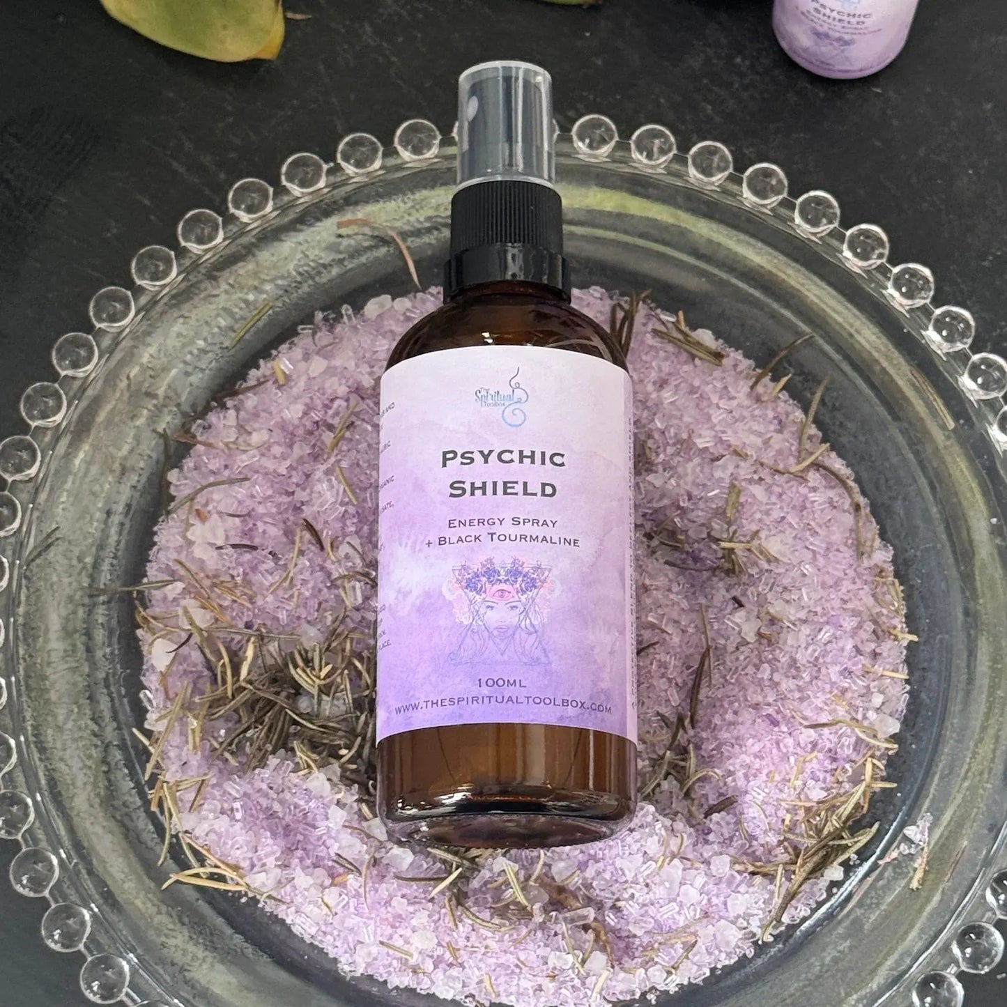 Bottle of 'Psychic Shield' energy spray on a decorative plate with purple sand and herbs.