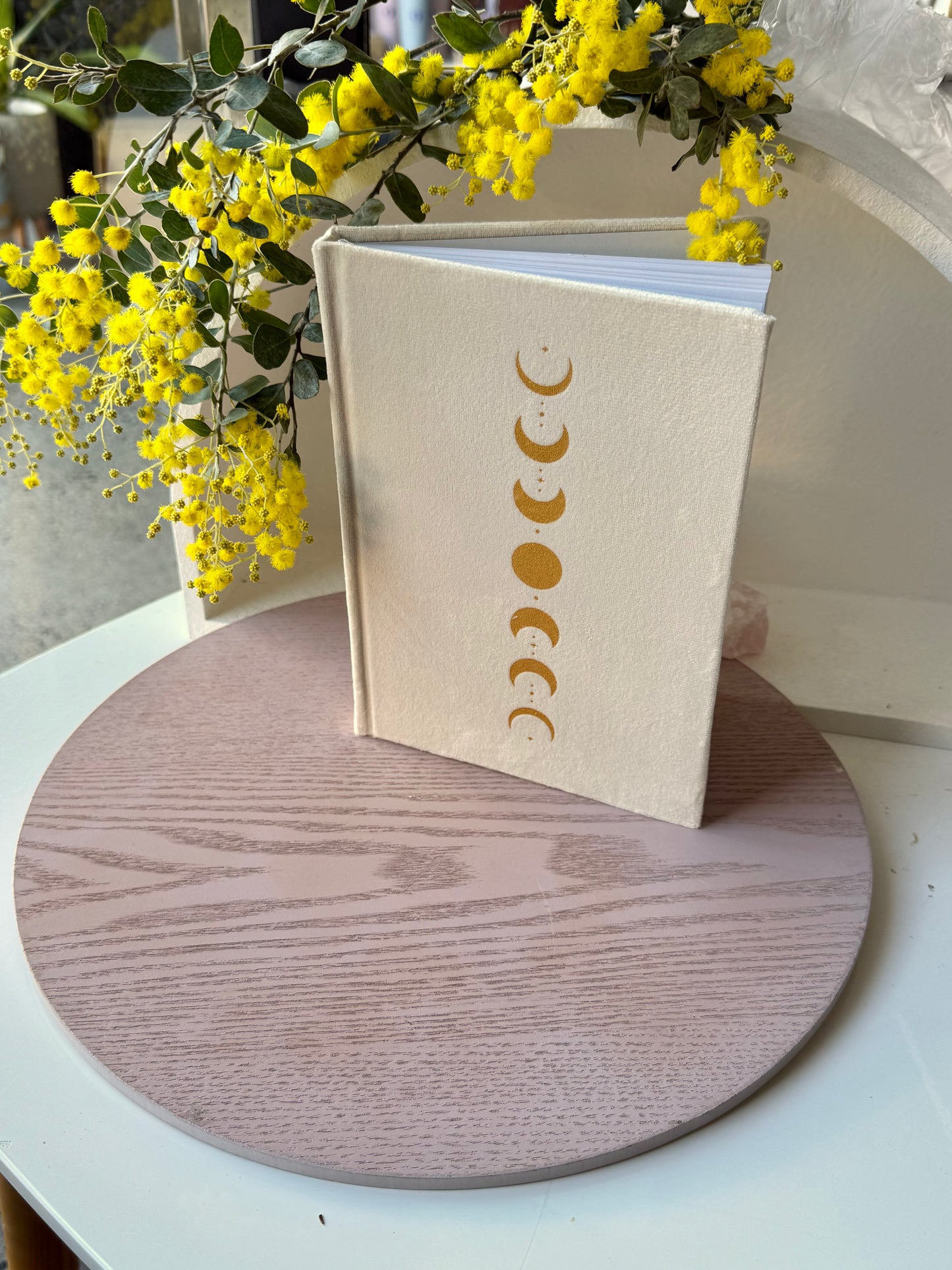 Beige notebook with gold moon phase design on a wooden surface with yellow flowers in the background