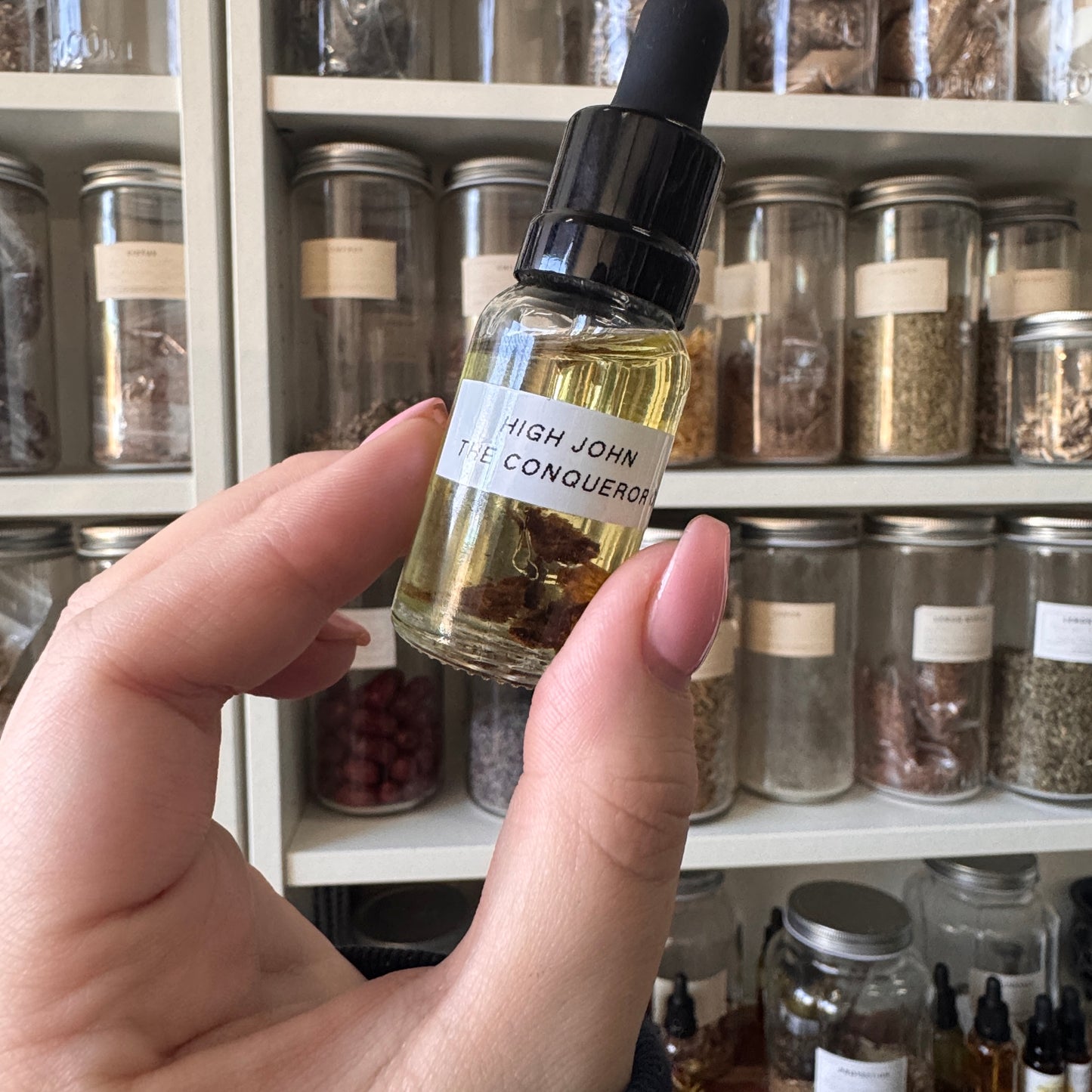 Hand holding a small bottle labeled 'High John the Conqueror' in front of shelves with jars.