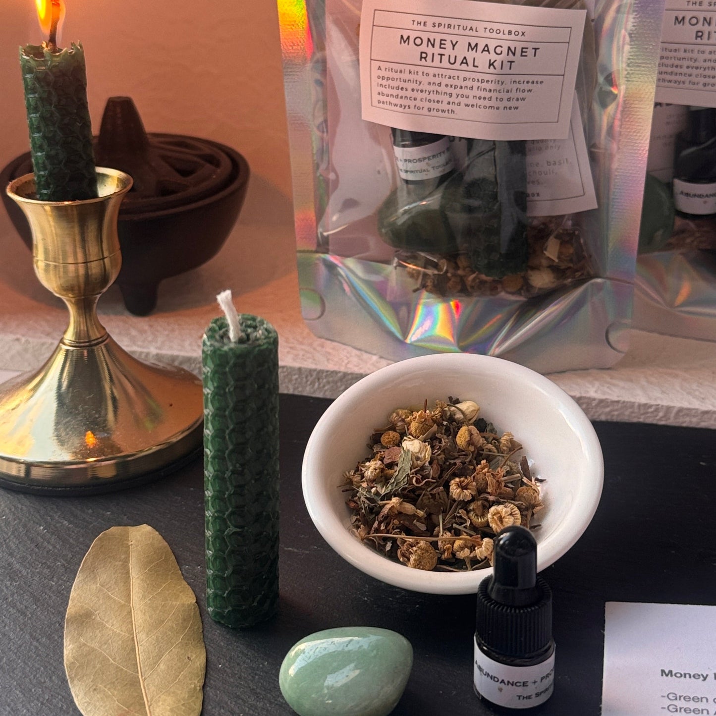 A ritual kit setup on a dark slate tray with a holographic pouch labeled 'Money Magnet Ritual Kit,' a small bowl of dried herbs, a green candle, a leaf, a stone, a dropper bottle, and a candle holder with a lit candle.