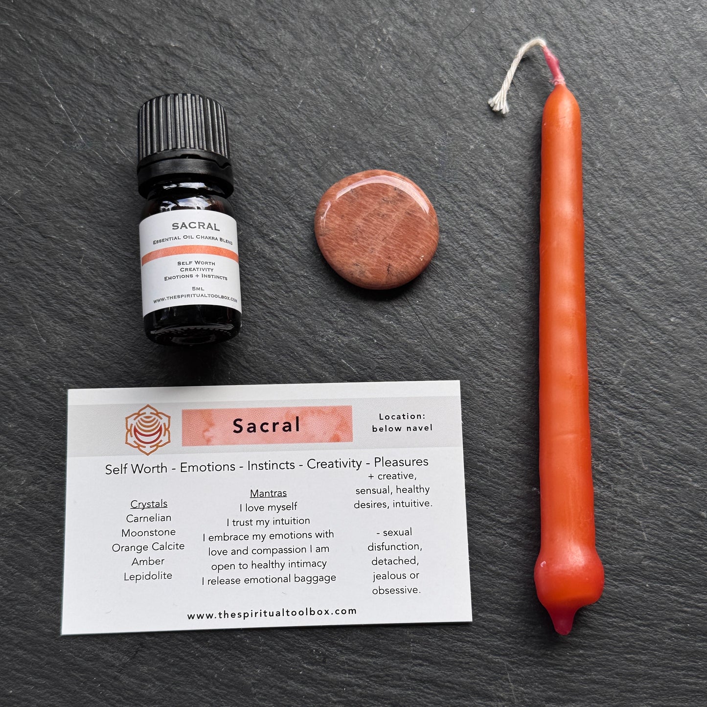 Sacral essential oil bottle, candle, crystal, and card on a dark surface