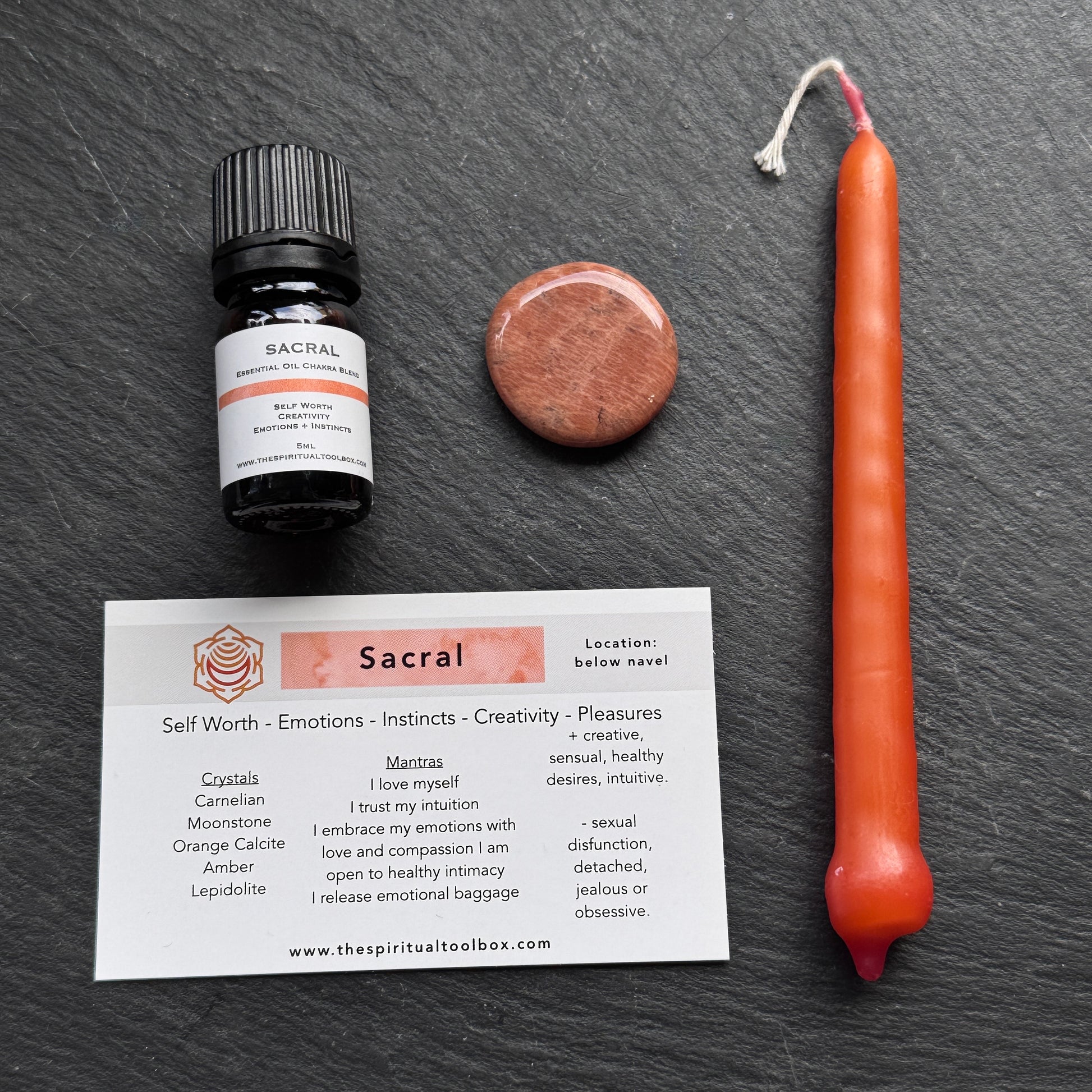 Sacral essential oil bottle, candle, crystal, and card on a dark surface