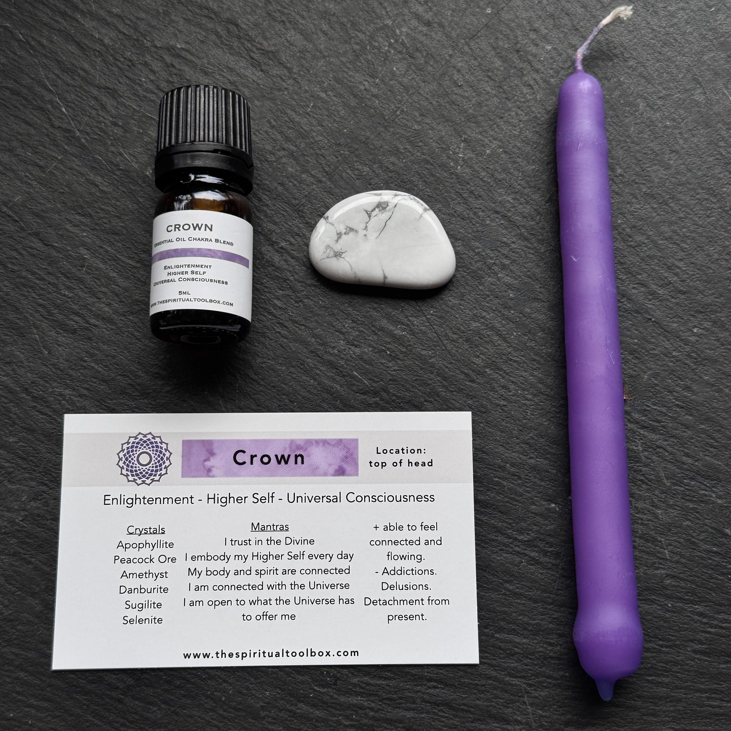 Crown chakra oil bottle, candle, stone, and card on a dark surface