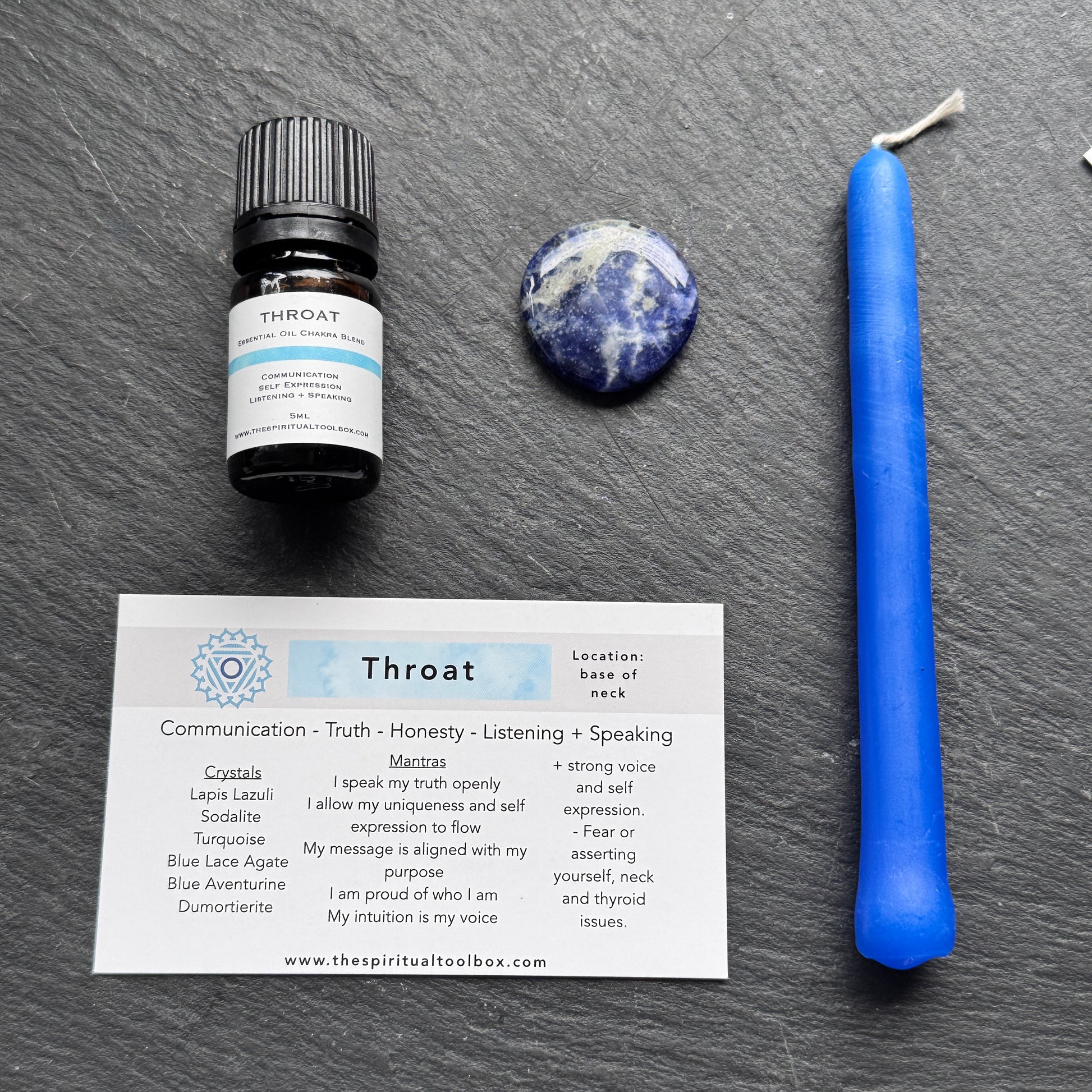 Throat chakra healing items including a bottle, candle, crystal, and card on a dark surface.