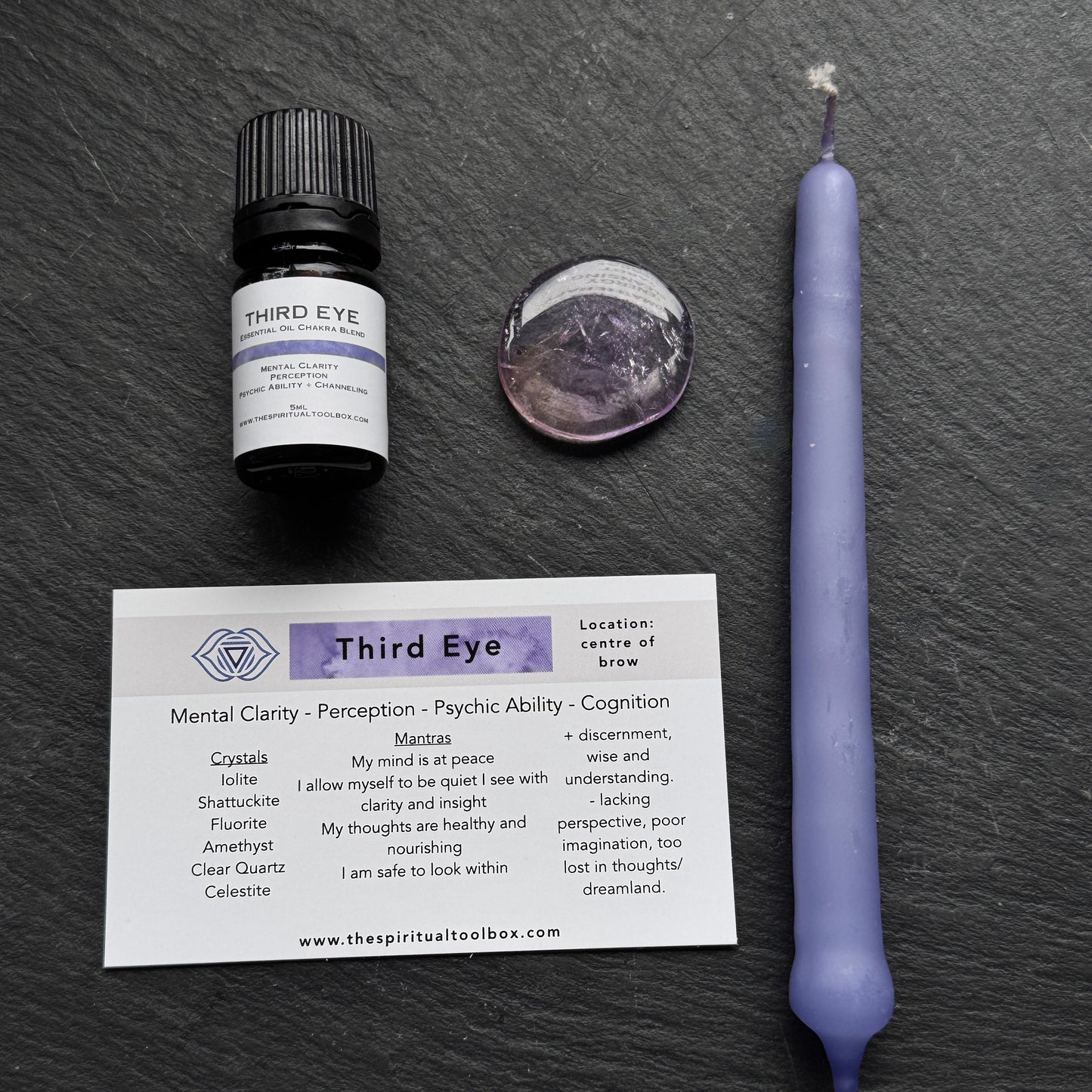 Vial labeled 'Third Eye', purple candle, amulet, and card on a dark surface