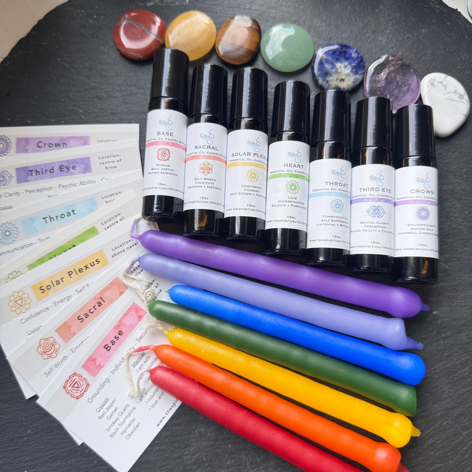 Set of essential oil bottles labeled for different chakras, candles in rainbow colors, and crystals on a dark surface.