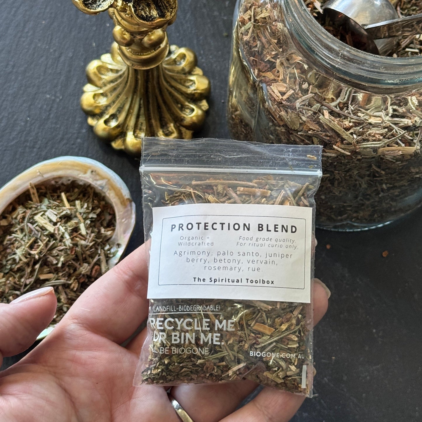 Hand holding a package of 'Protection Blend' with a lit candle and jars in the background.
