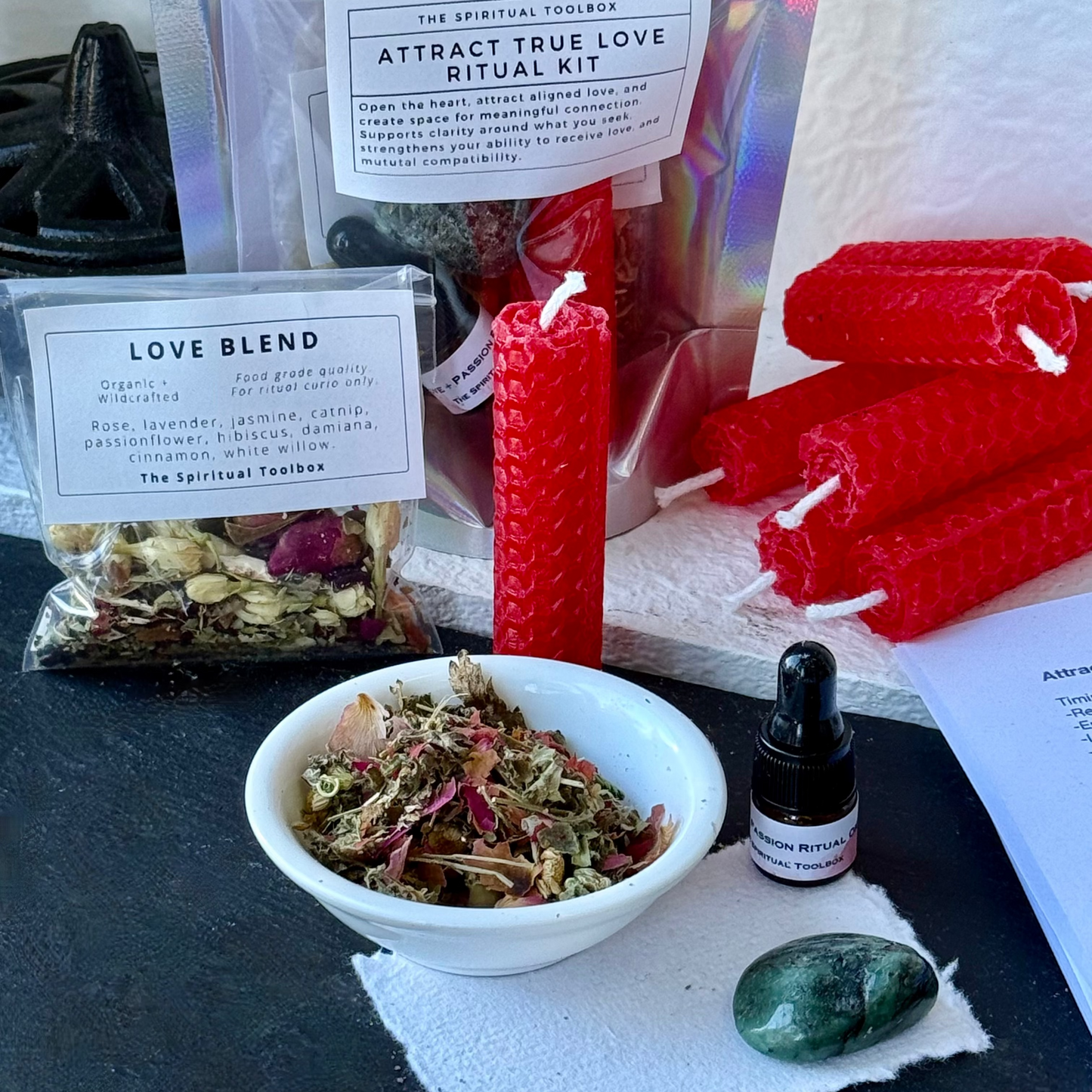 Attract True Love Ritual Kit by The Spiritual Toolbox, featuring red beeswax candles, a bowl of dried herbs, a small dropper bottle, a green crystal, and a bag of rose petals, arranged on a dark surface.