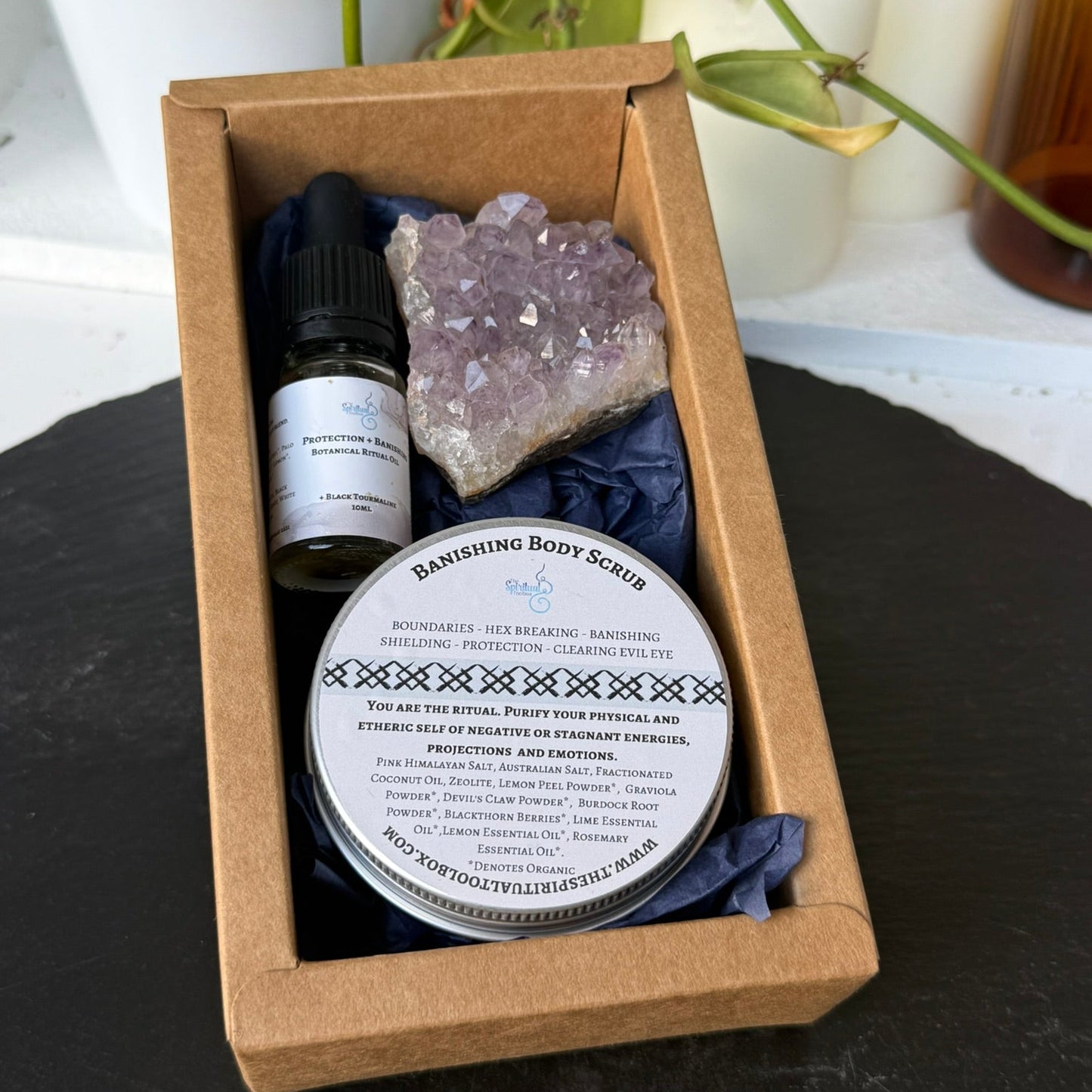 Box containing a bottle, a crystal, and a container labeled 'Banishing Body Scrub' on a dark surface with plants in the background.