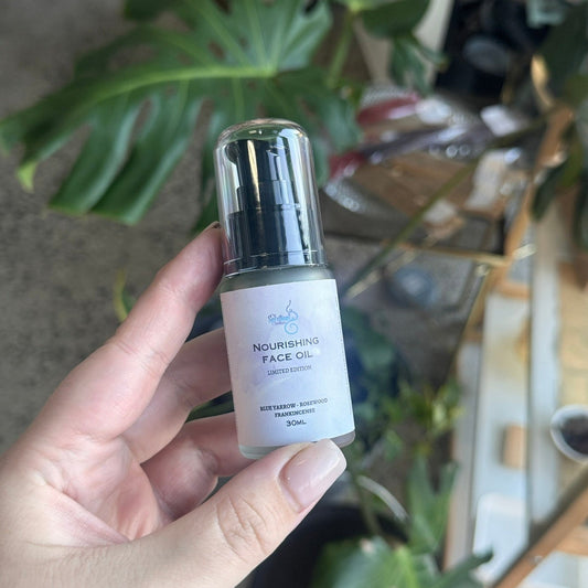 A hand holding a small bottle of Nourishing Face Oil with a clear cap and white label, set against a blurred indoor background with green plants and shelves.