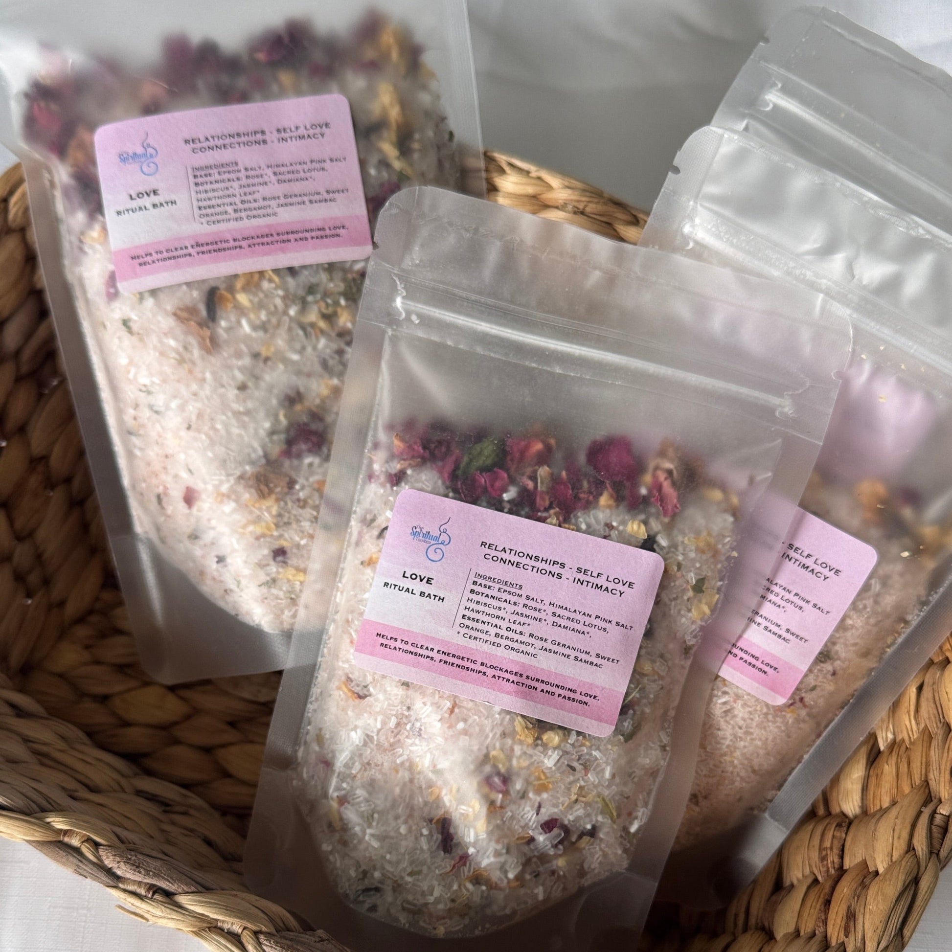 Three packages of bath salts with pink labels in a woven basket on a white background