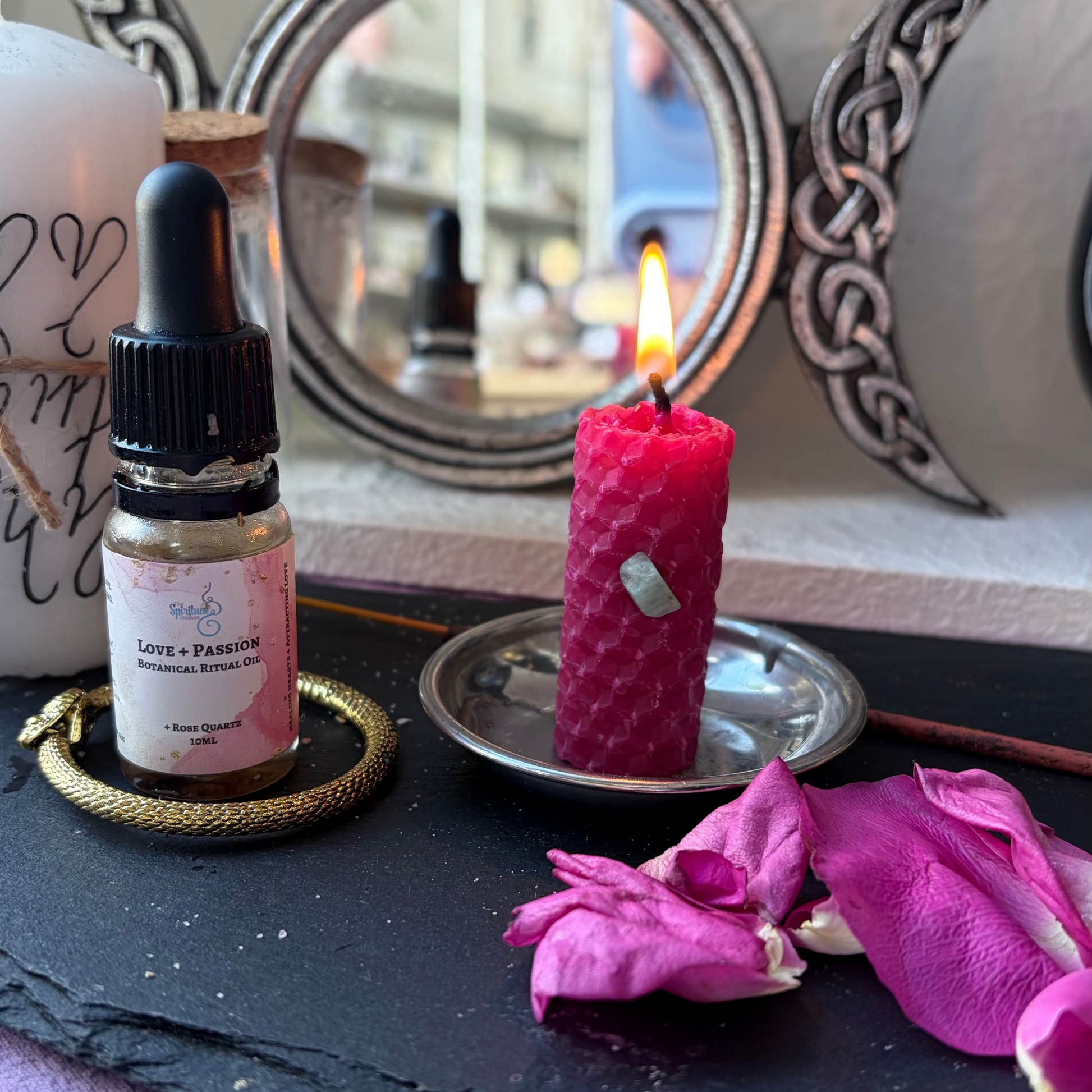 A small glass dropper bottle labeled 'Love & Passion' with a black dropper cap, styled on a black slate tray beside a pink candle, pink petals, and a white candle in a decorative setting.