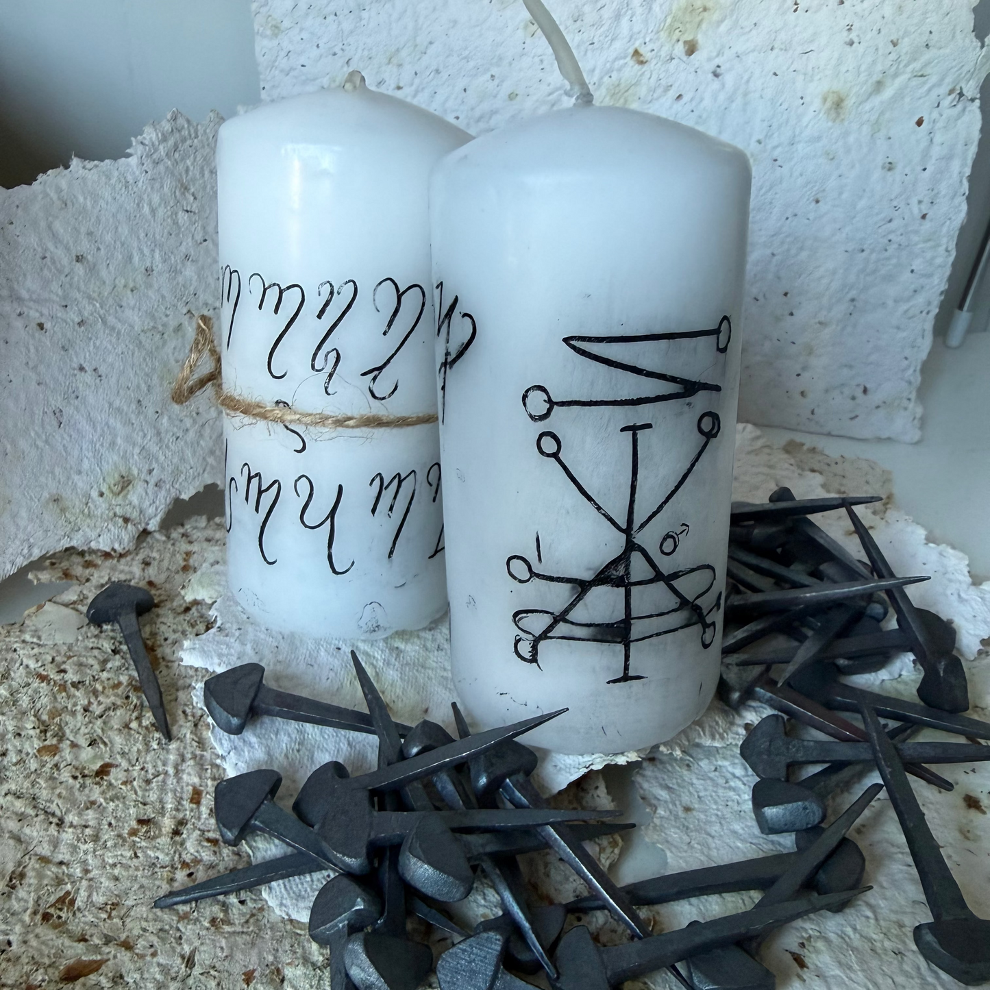 Two white pillar candles with black occult-style symbols and runic lettering, displayed on a rustic surface with scattered dark metal nails and torn white paper.
