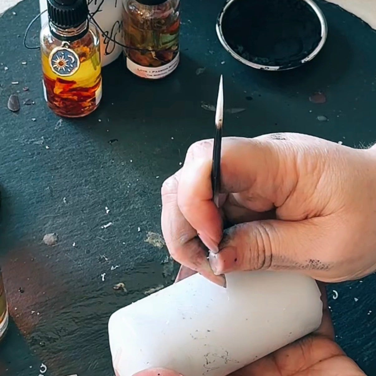 A close-up of hands carving a white cylindrical candle wax pillar using a small carving tool, with various wax bottles and a black tray in the background.