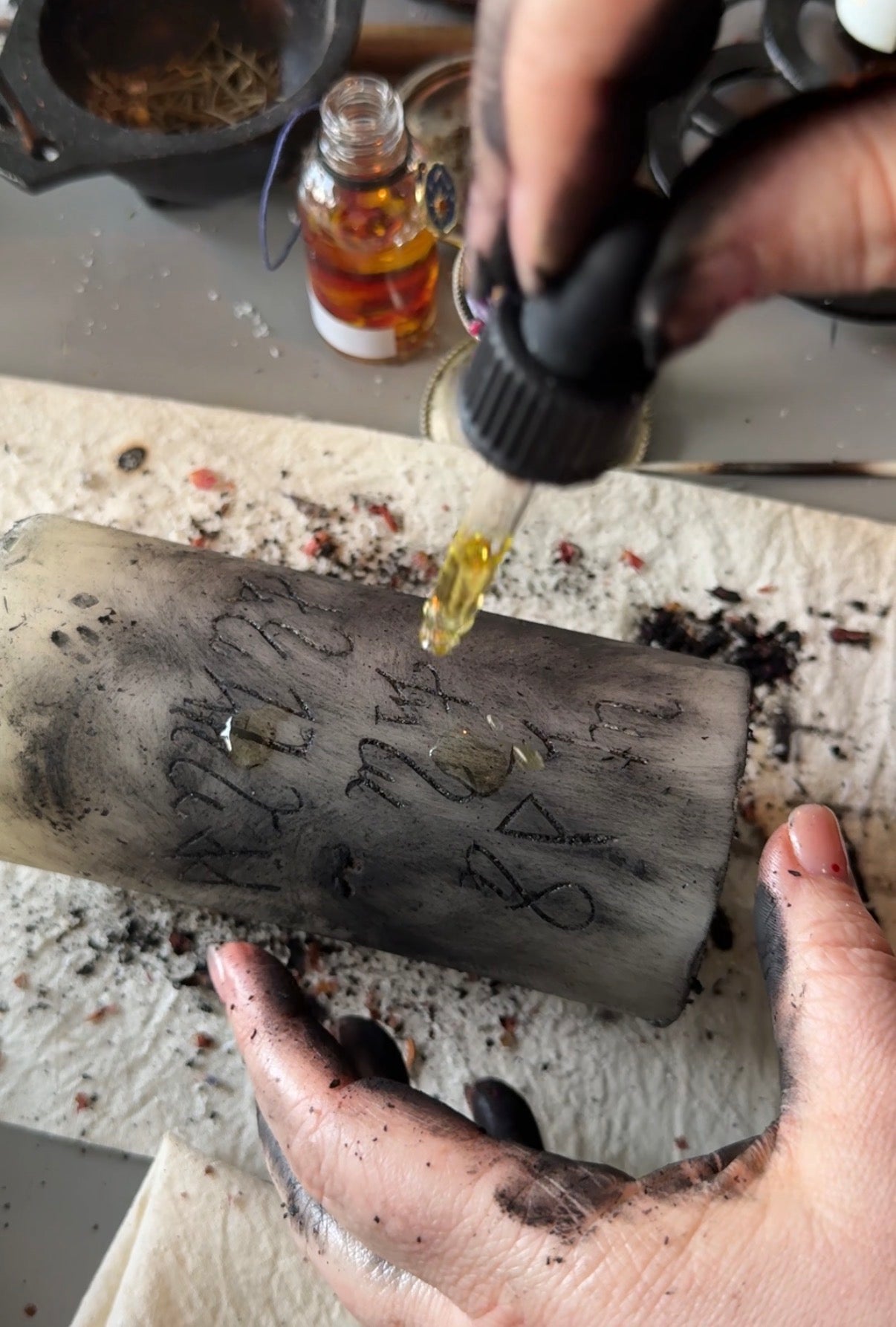 A close-up of hands applying a golden liquid from a dropper onto a dark, rectangular candle or stone slab marked with symbols, with a small amber bottle and workshop items in the background.