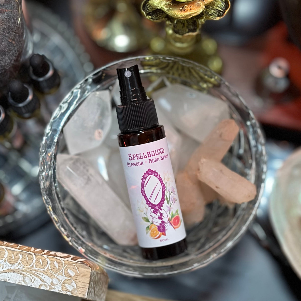 A small amber spray bottle labeled 'SPELLBOUND' sits in a glass bowl filled with ice and crystals, surrounded by candles, crystals, and other spa-like props.