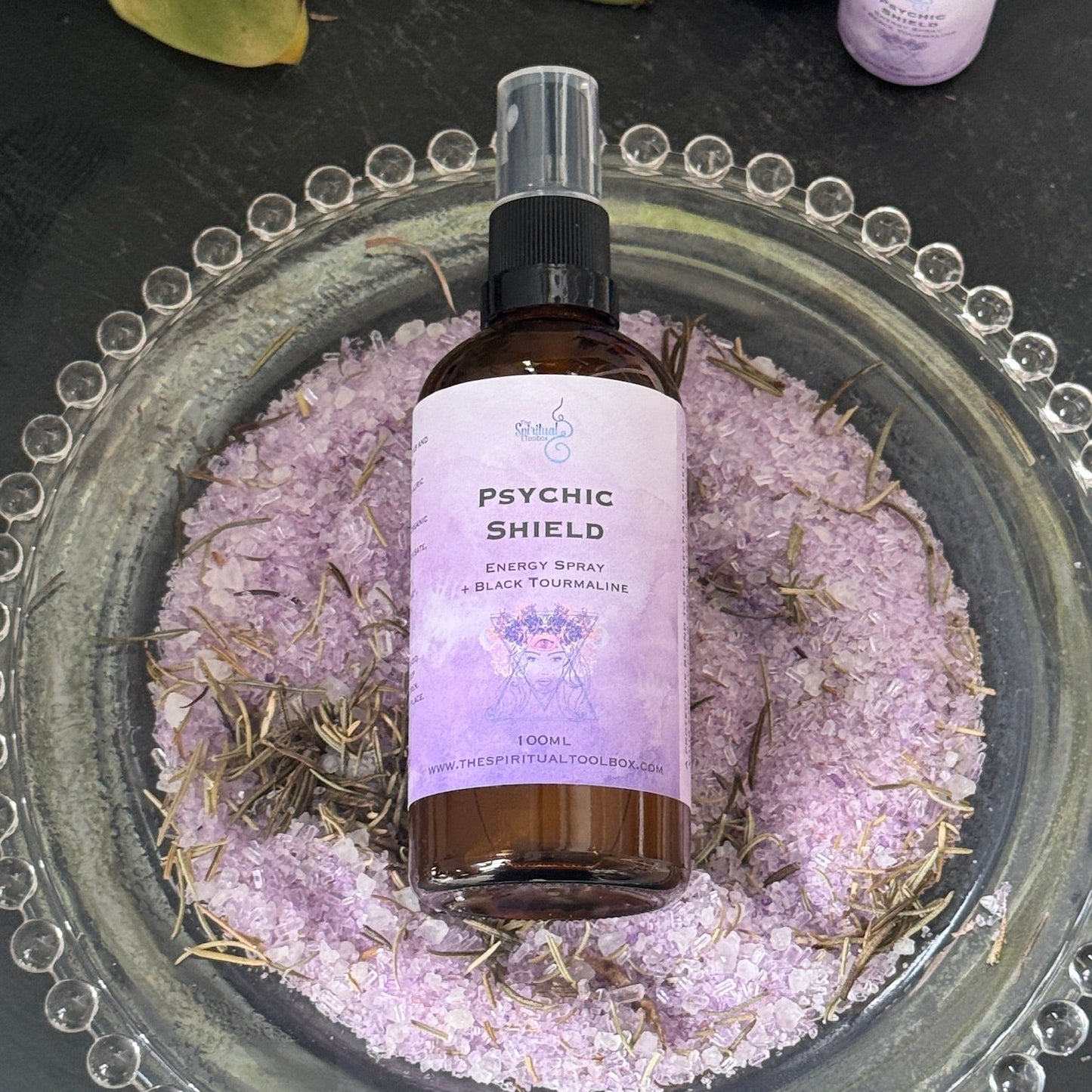 Bottle of 'Psychic Shield' energy spray on a decorative plate with purple sand and herbs.
