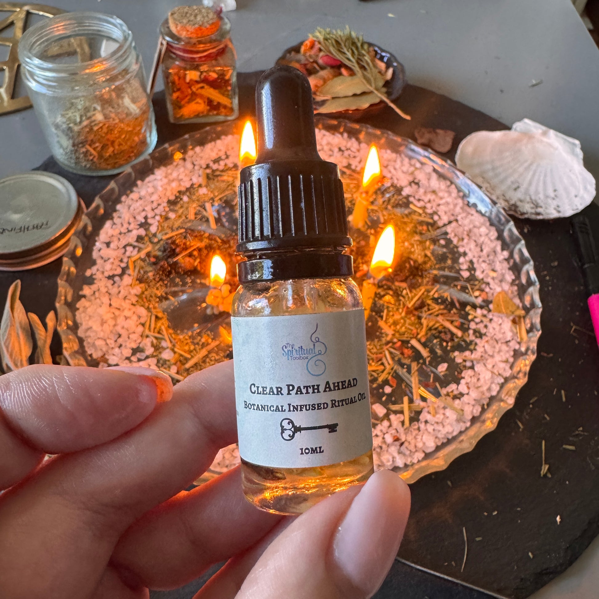 A hand holds a small glass dropper bottle labeled 'Clear Path Ahead Botanical Infused Ritual Oil' in front of a ritual setup with lit candles, crystals, dried herbs, and a shell.