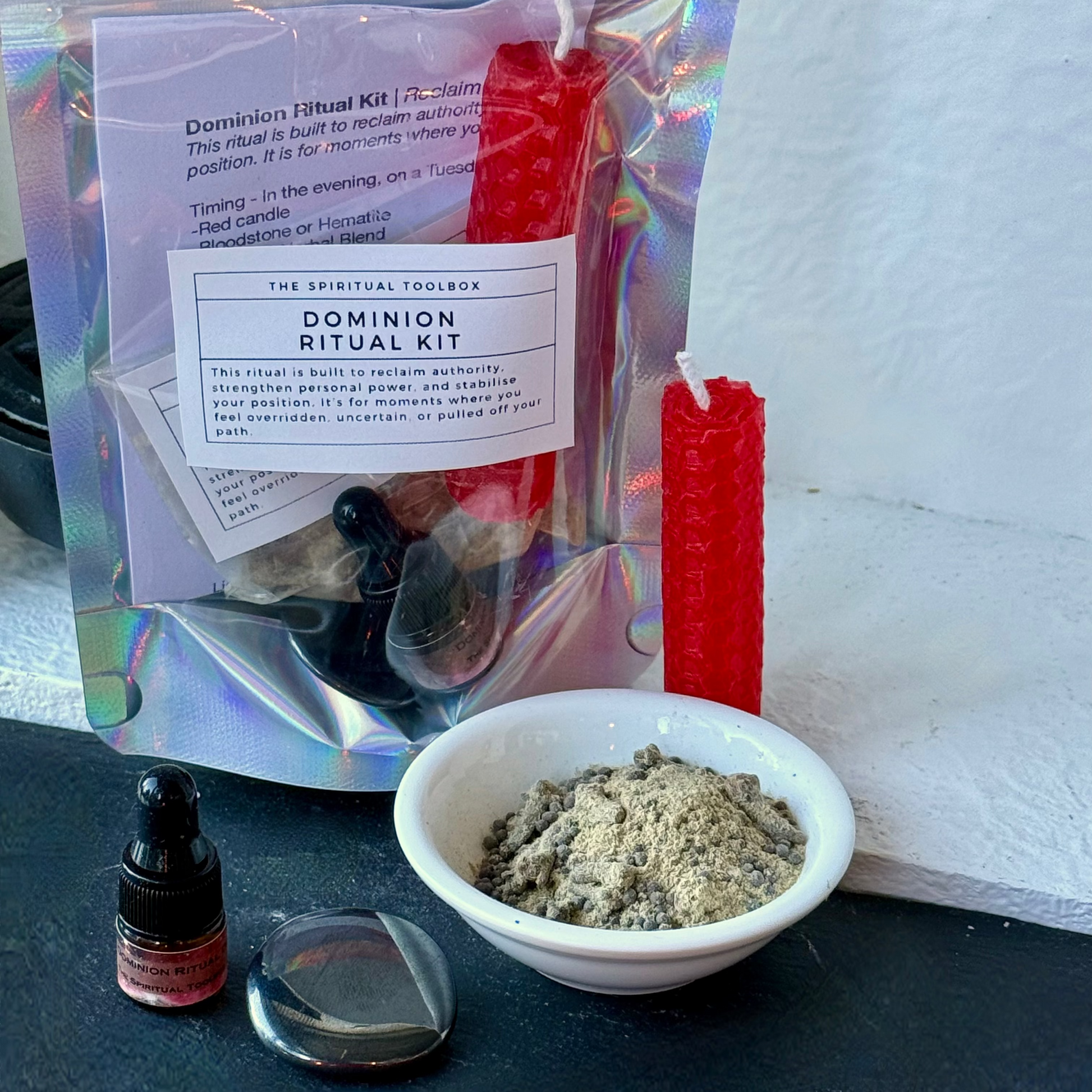 A Dominion Ritual Kit displayed on a dark surface against a white wall. The kit includes a holographic pouch with a white label, a red taper candle, a small dropper bottle, a bowl of gray powder, and a clear glass dish.