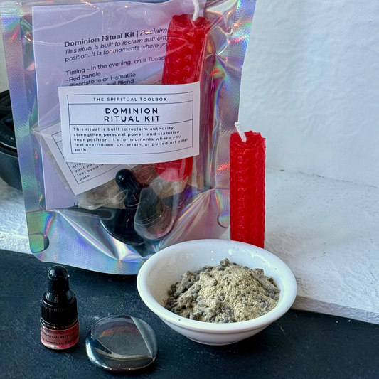 A Dominion Ritual Kit displayed on a dark surface against a white wall. The kit includes a holographic pouch with a white label, a red taper candle, a small dropper bottle, a bowl of gray powder, and a clear glass dish.
