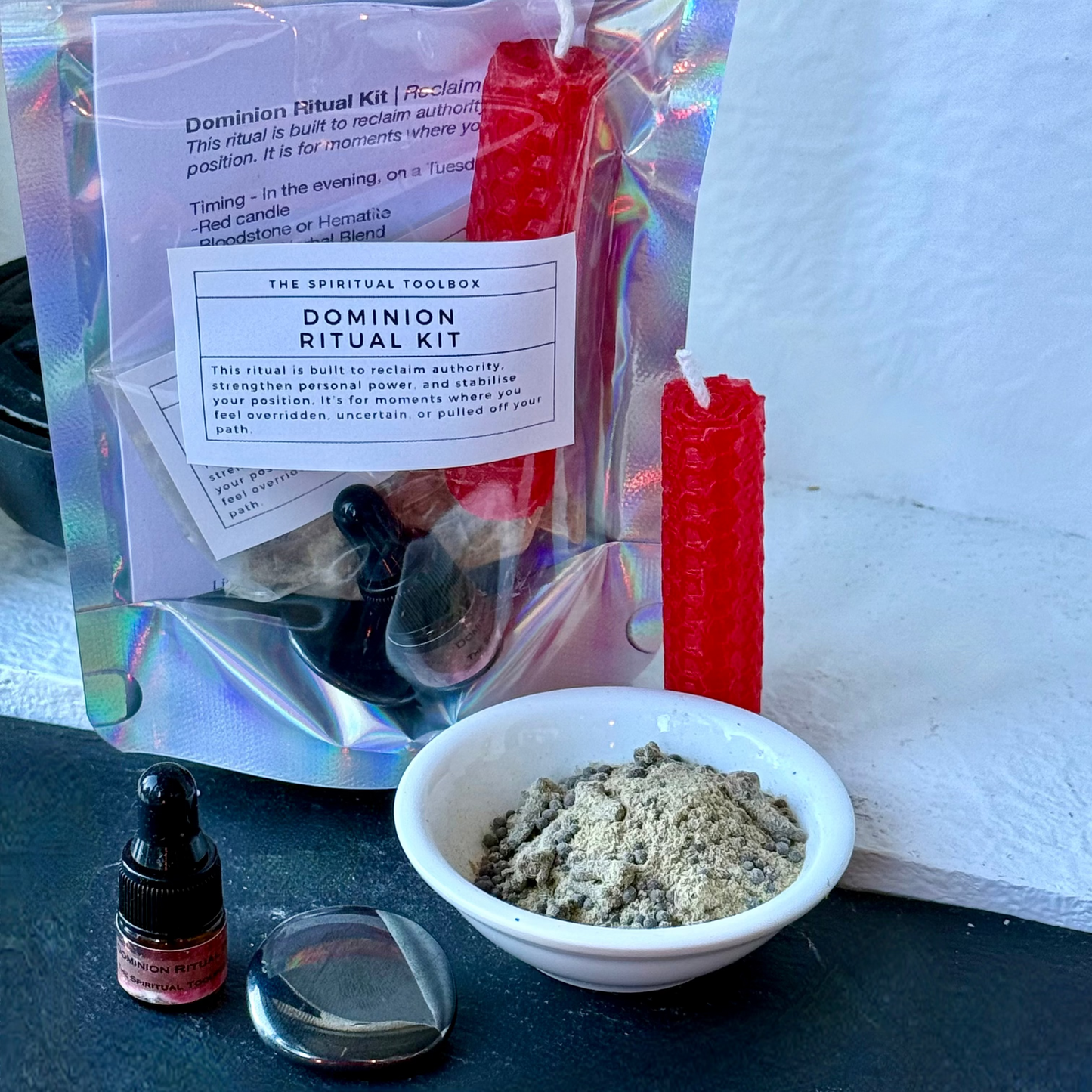 A Domination Ritual Kit by The Spiritual Toolbox displayed on a black surface against a white wall. The kit includes a holographic pouch with a white label, red wax candles, a small white bowl filled with gray powder, a glass dropper bottle, and a clear glass container.