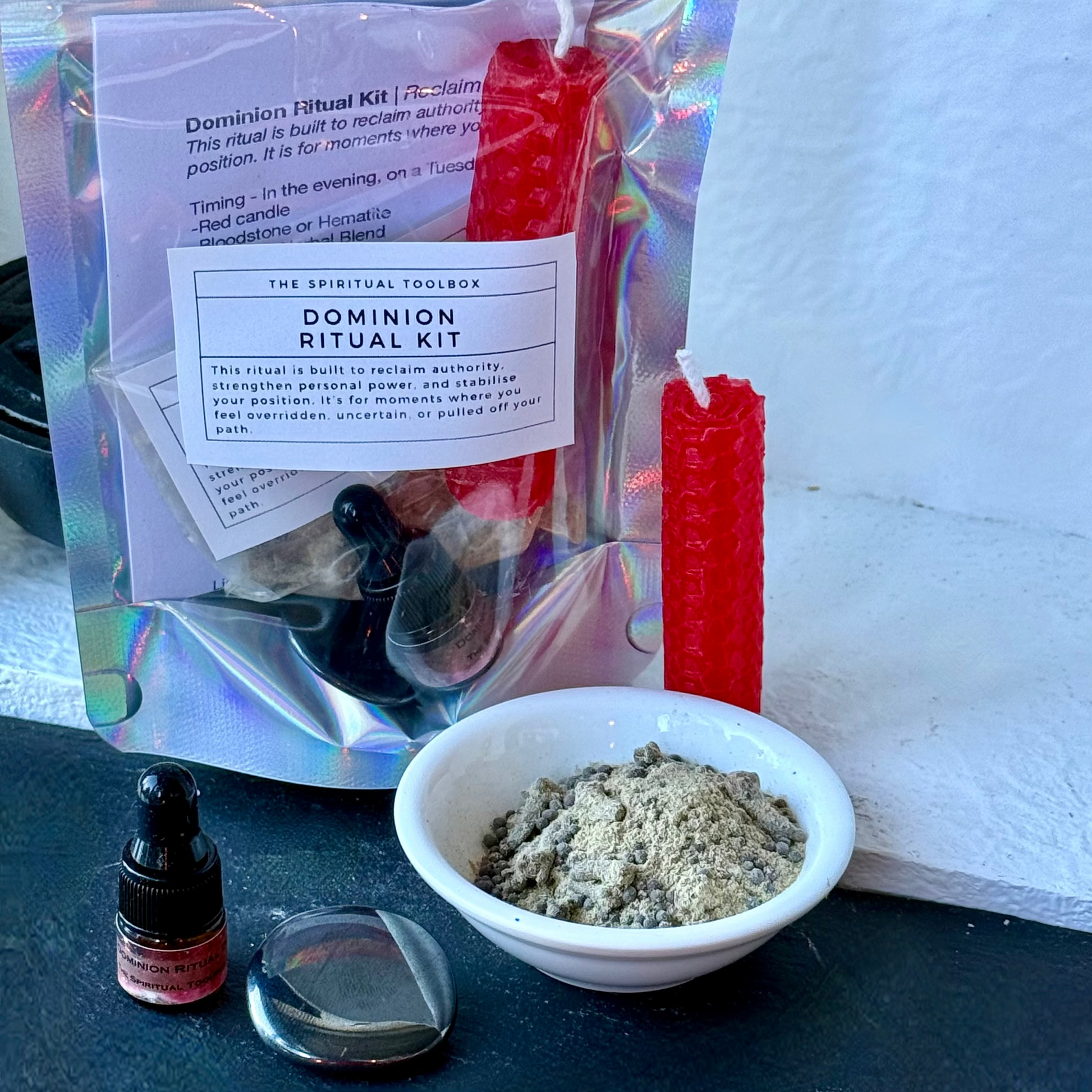 A Domination Ritual Kit by The Spiritual Toolbox displayed on a black surface against a white wall. The kit includes a holographic pouch with a white label, red wax candles, a small white bowl filled with gray powder, a glass dropper bottle, and a clear glass container.