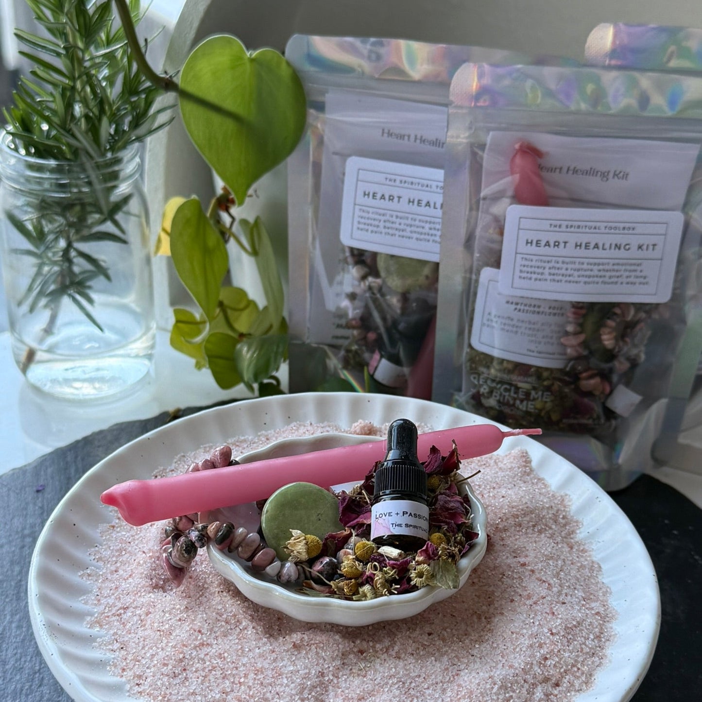 Heart healing kit with candle, bottle, and bowl on a white plate with plants in the background.