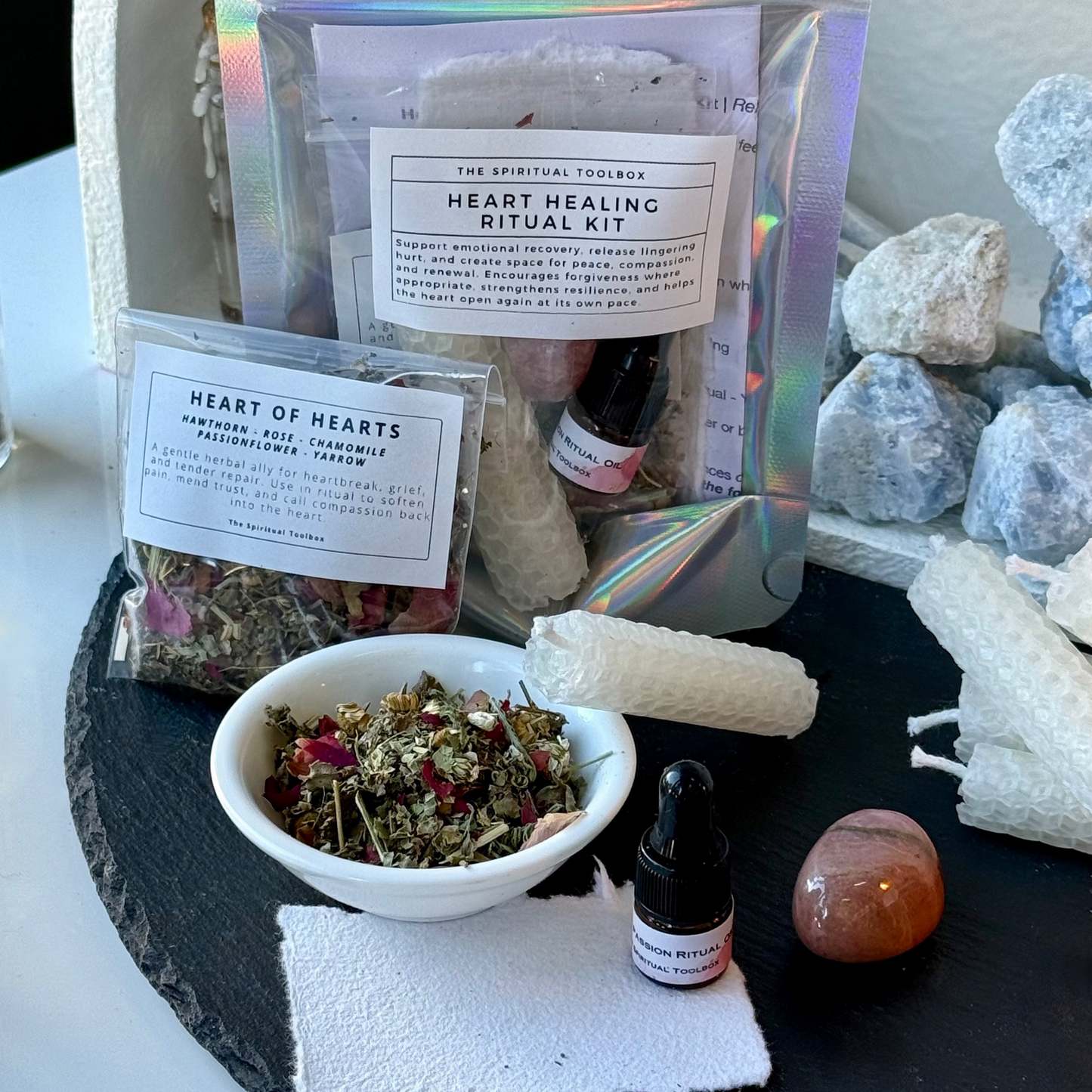 Heart Healing Ritual Kit by The Spiritful Foolbox, featuring an iridescent stand-up pouch, a smaller packet of dried herbs, a white bowl of loose blend, a small dropper bottle, and ritual tools arranged on a black tray with crystals.