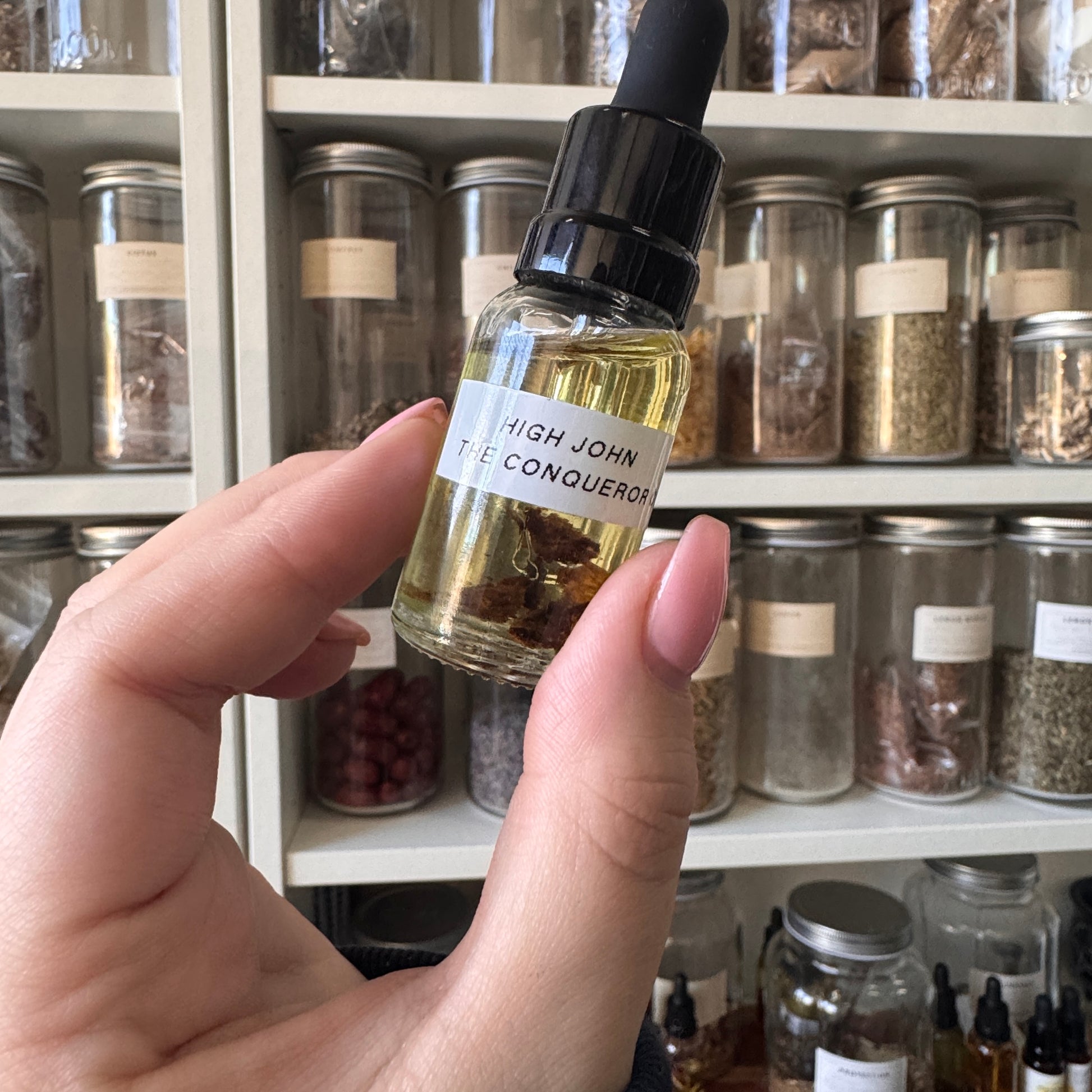 Hand holding a small bottle labeled 'High John the Conqueror' in front of shelves with jars.