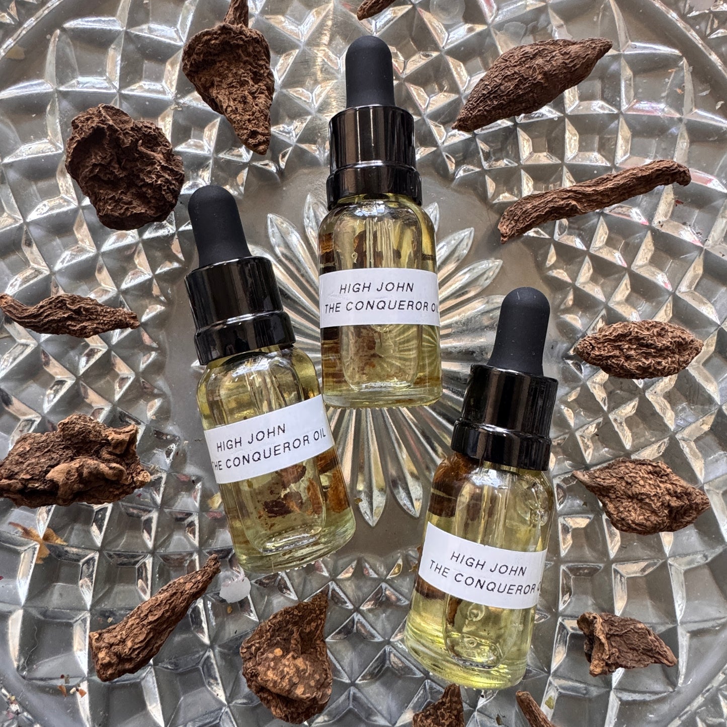 Three bottles of 'High John the Conqueror' oil with droppers on a textured metallic surface.