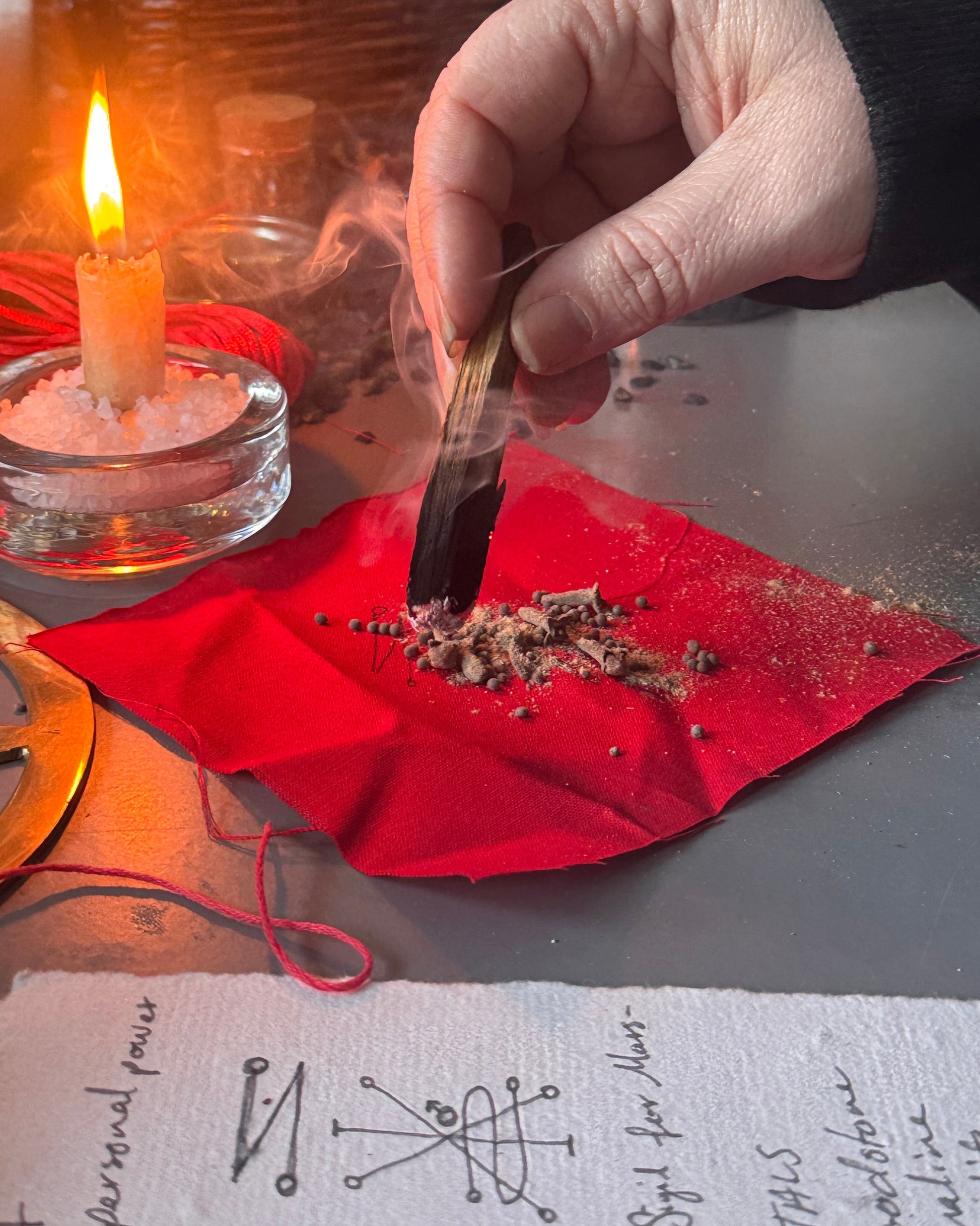A hand holds a burning incense stick over a red paper square on a tabletop, with a lit candle in a glass holder nearby and blurred ritual or decorative objects in the background.