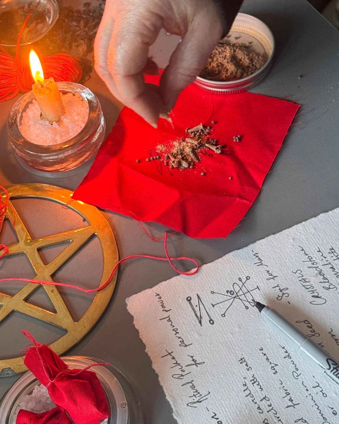 A close-up of a hand sprinkling herbs onto a red cloth on a tabletop, surrounded by candles, a pentagram symbol, a handwritten note, and ritual items.