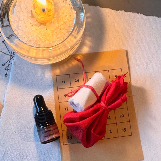 A ritual or altar setup featuring a lit candle in a glass holder, a small dropper bottle, a brown parchment calendar grid, a rolled white scroll tied with red string, and a red fabric pouch on a white cloth.