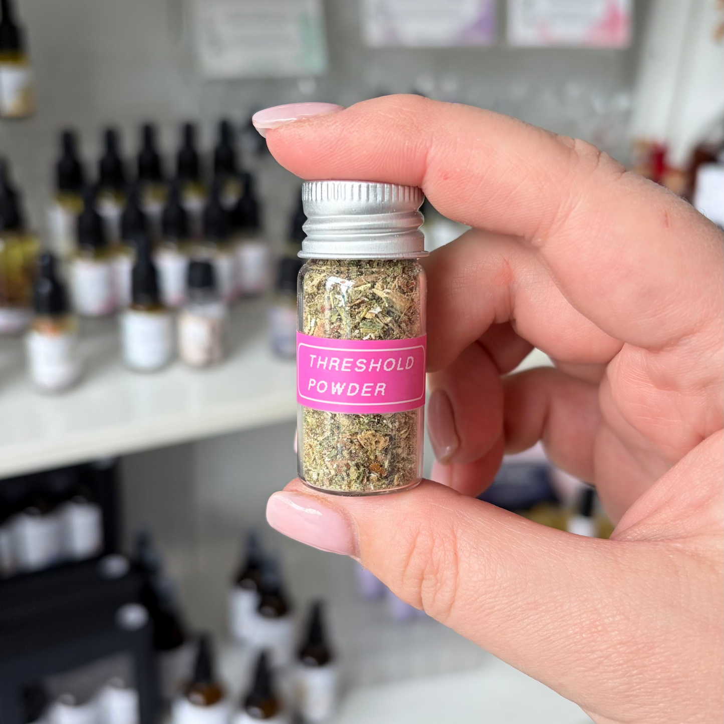 A hand holding a small glass vial of 'Thresholdic Powder' with a pink label and silver cap, set against a blurred background of shelves with other bottles.