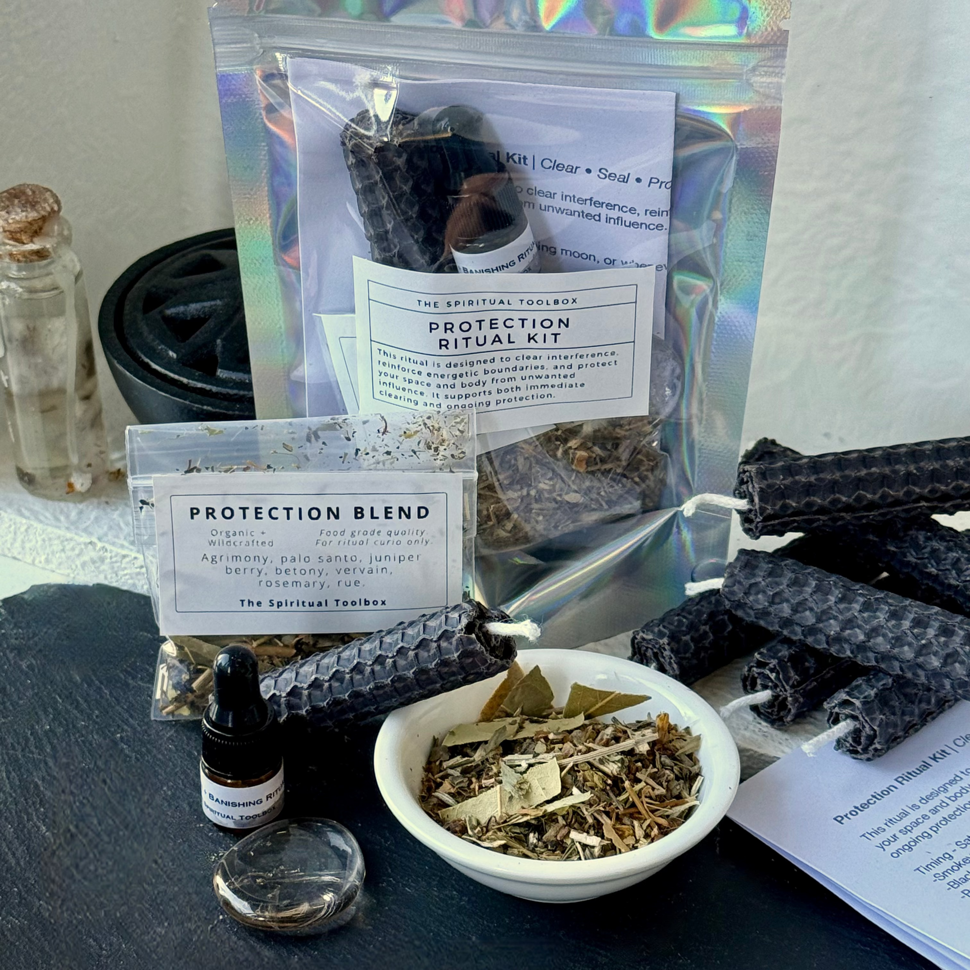 Protection Ritual Kit by The Sacred Turmeric, featuring a holographic pouch of protection blend, a small dropper bottle, a bowl of dried herbs, beeswax candles, and a crystal ball on a dark slate surface.