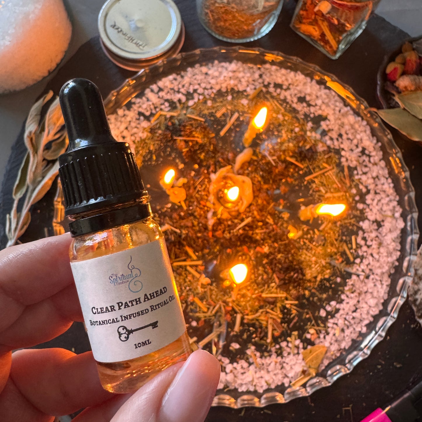 A hand holds a small glass dropper bottle labeled 'Wildberry Clear Path Ahead' in front of a glass bowl filled with dried herbs and lit candles.