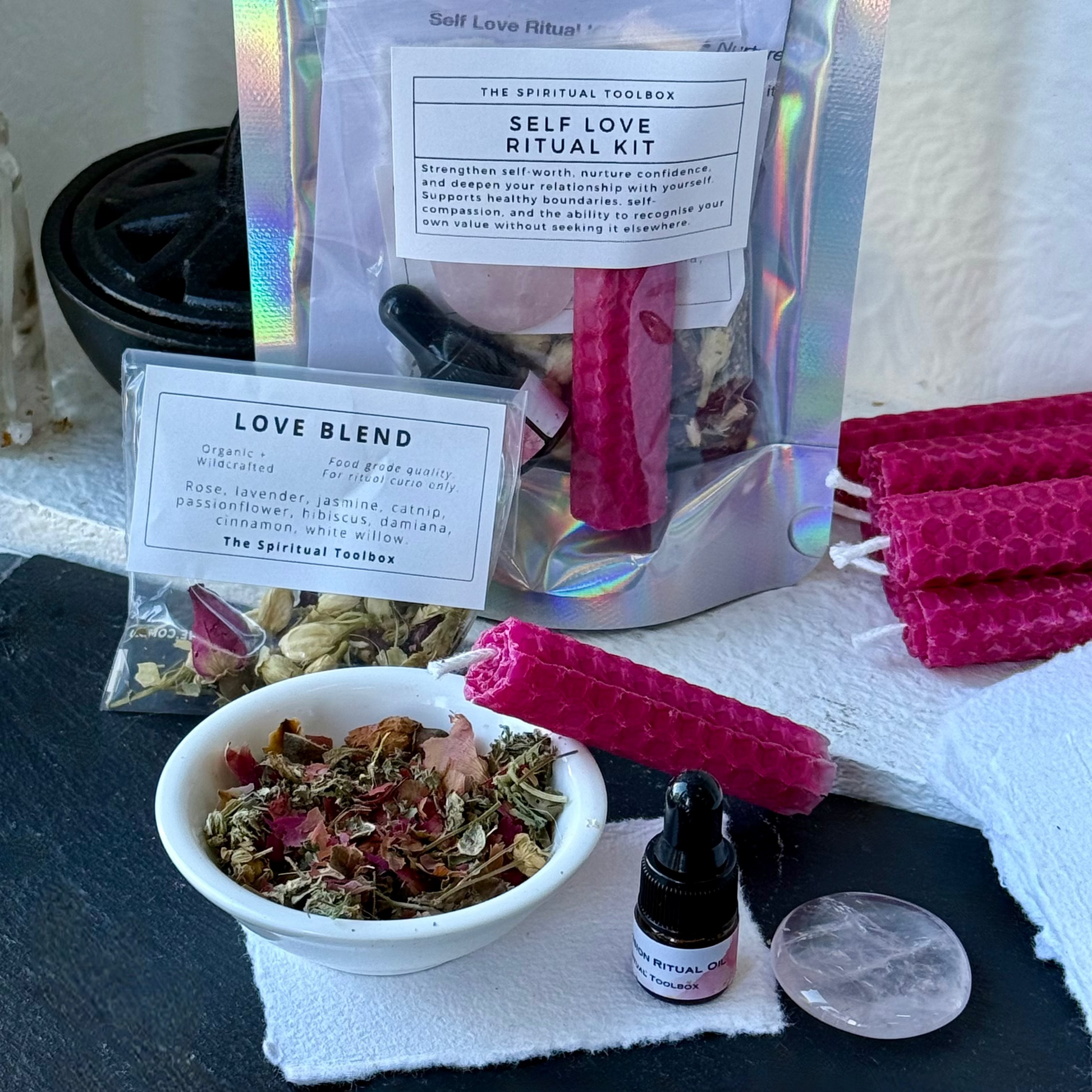Self Love Ritual Kit by The Spiritual Toolbox, featuring a holographic stand-up pouch with a pink wax candle, a small white bowl of dried herbs and flowers, a pink honeycomb wax candle, a small amber dropper bottle, and a clear crystal, arranged on a dark slate surface with a white cloth.