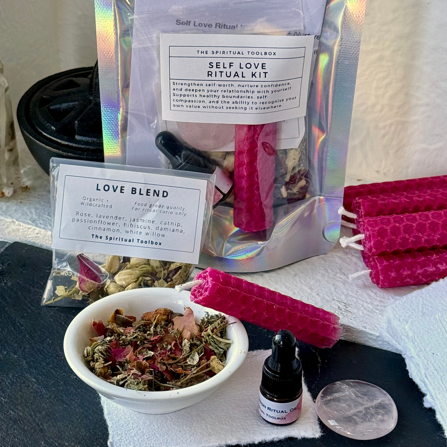 Self Love Ritual Kit by The Spiritual Toolbox, featuring a holographic stand-up pouch with a 'Self Love Ritual Kit' label, a 'Love Blend' herbal sachet, pink beeswax candles, a small dropper bottle, and a clear crystal on a white cloth over a dark surface.